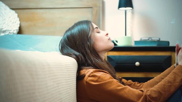 Young Hispanic woman sitting on the floor beside her bed, enjoying a peaceful and quiet moment to think, reflect, and reconnect with herself at home.