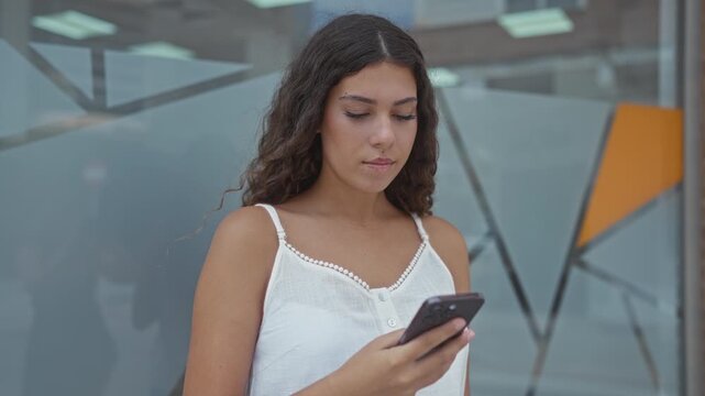 Young woman holding smartphone and checking screen while leaning against geometric glass building facade, wearing white camisole and neutral expression; serene.