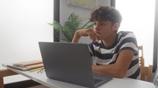 Teenage boy pointing finger at laptop screen and typing at desk in building; concentration learning.