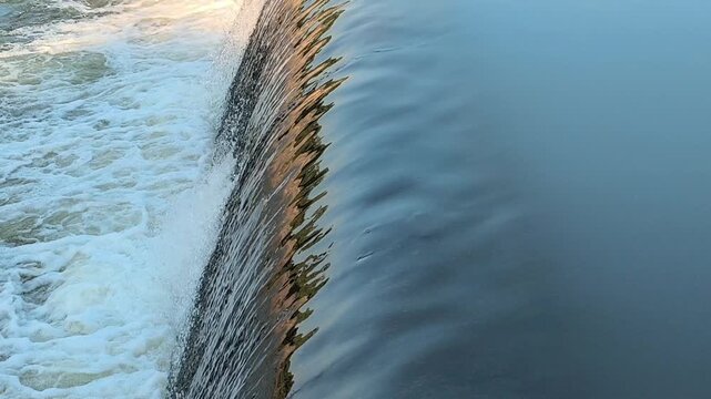 Fast-flowing water rushing over a small dam structure, creating white foam, turbulent currents, and dynamic cascades that highlight the power and movement of flowing water