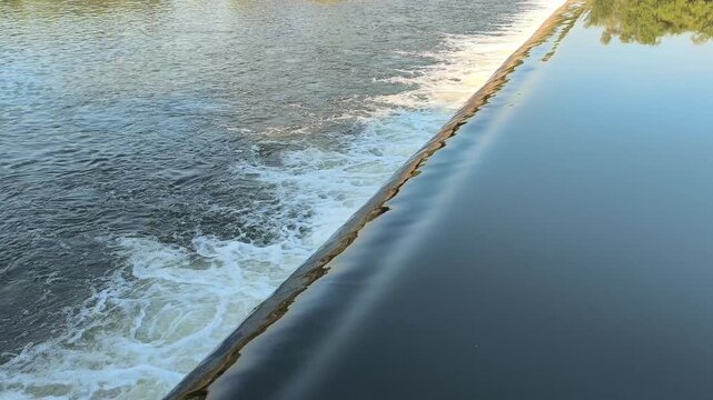 Fast-flowing water rushing over a small dam structure, creating white foam, turbulent currents, and dynamic cascades that highlight the power and movement of flowing water