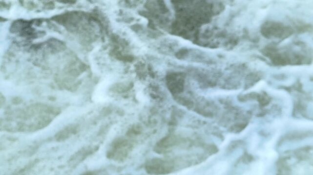 A blurred and defocused view of turbulent water swirling, churning, and producing soft foamy patterns, creating an abstract fluid background with dynamic motion and atmospheric texture.
