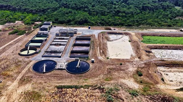 Aerial parallax reveals circular clarifiers and treatment basins at wastewater facility surrounded by forest near Plage des Oceanides