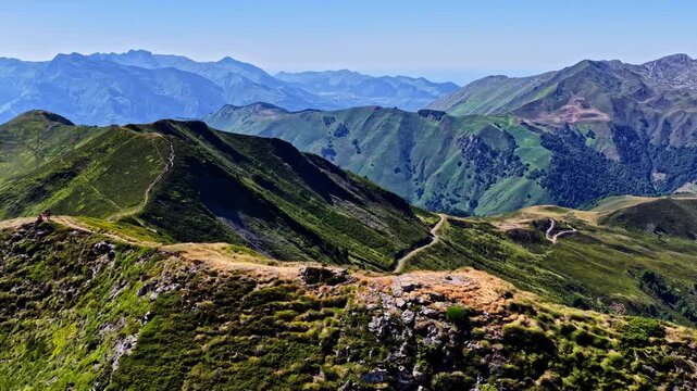 Aerial parallax follows two tiny hikers along winding ridge trail with layered green peaks and distant rocky summits behind at Col d Aubisque, France