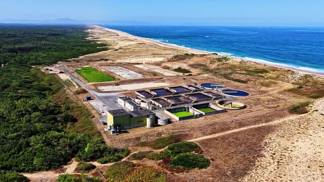Aerial orbit reveals coastal water treatment facility with circular tanks sitting between pine forest and Plage des Oceanides shoreline