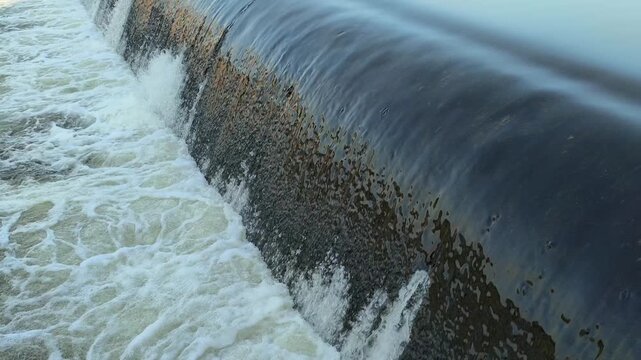 Fast-flowing water rushing over a small dam structure, creating white foam, turbulent currents, and dynamic cascades that highlight the power and movement of flowing water