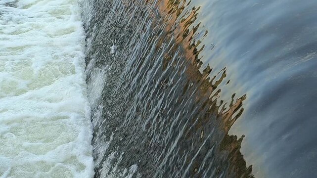 Fast-flowing water rushing over a small dam structure, creating white foam, turbulent currents, and dynamic cascades that highlight the power and movement of flowing water