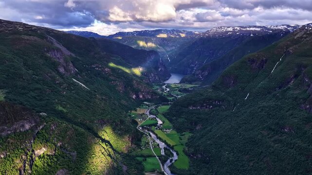 Aerial timelapse shows patches of sunlight traveling across green Aurland valley with winding river toward Aurlandsfjord Norway