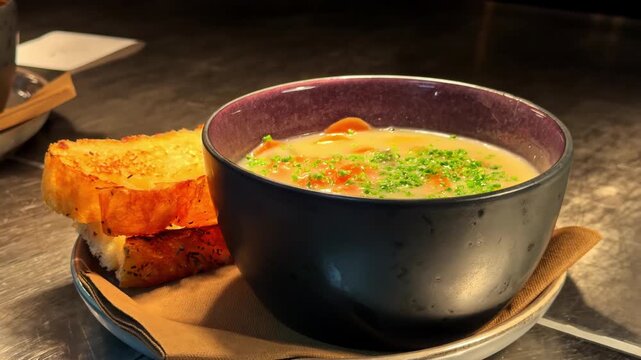 Steaming bowl of creamy carrot soup topped with chives served beside toasted garlic bread on rustic restaurant table in Norway