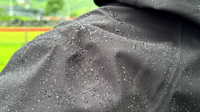 Close up slow motion shot shows raindrops beading on waterproof black raincoat fabric with green field blurred in background