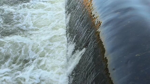 Fast-flowing water rushing over a small dam structure, creating white foam, turbulent currents, and dynamic cascades that highlight the power and movement of flowing water