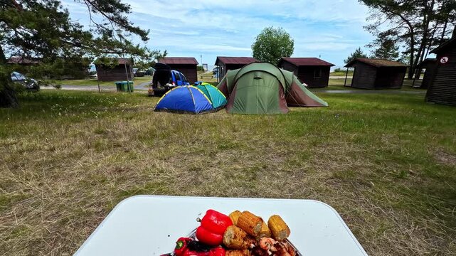 Camera tilts down and zooms into grilled lobster seafood platter with shrimp, corn and peppers on foil tray at campsite surrounded by tents, cabins and pine trees
