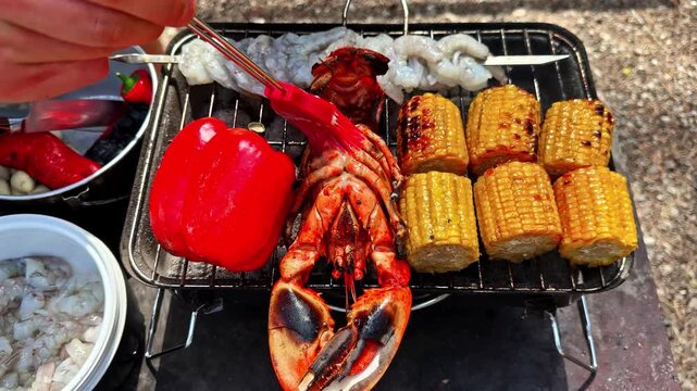 Hand brushes grilled lobster tail with bbq sauce on portable grill beside shrimp skewers, corn on cob and red bell pepper