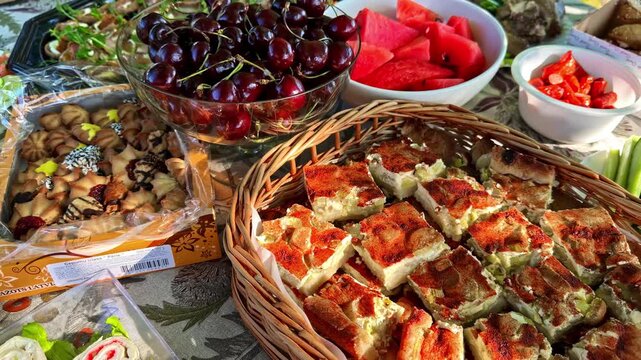 Outdoor buffet table displays fresh cherries watermelon roasted potatoes sliced meat sausages and pickles on floral tablecloth
