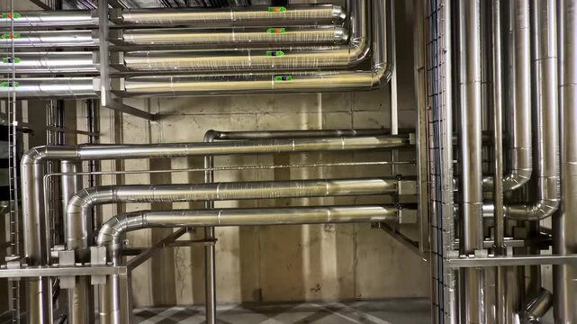Camera moves downward through rows of stainless steel pipes and ductwork inside industrial mechanical room with concrete walls