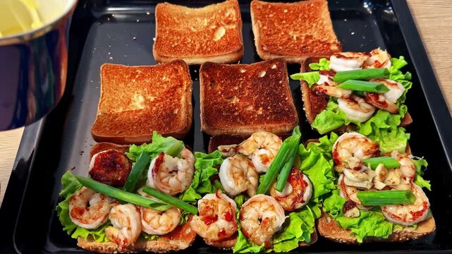 Hand uses tongs to add creamy sauce onto shrimp toast sandwiches topped with lettuce and green onion on black tray