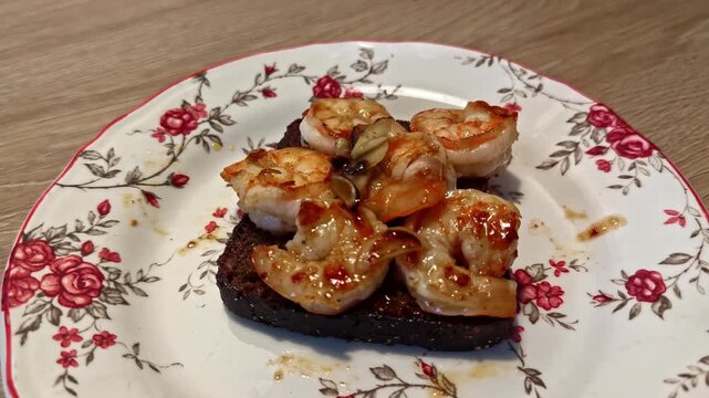 Orbit close up shows grilled garlic shrimp plated on toasted dark bread with vintage rose patterned china dish on wooden table