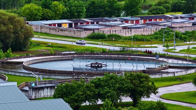 Circular wastewater treatment clarifier basin sits beside a road and green trees on an overcast day, viewed from above.