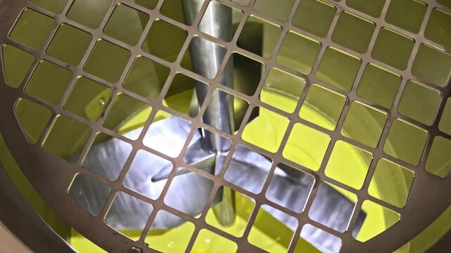 Yellow liquid swirls around metal blades inside a covered industrial agitator, viewed through the protective grid.