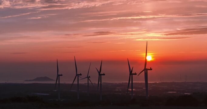 Timelapse of sun setting behind row of wind turbines along Jeju coastline with slow zoom and vivid orange sky