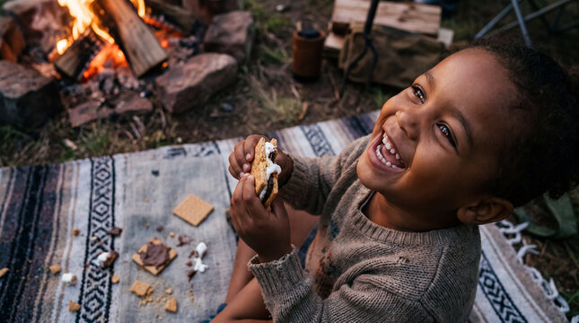 A smiling toddler looks up at the camera while holding a gooey s'more near a campfire for National S'mores Day lifestyle marketing