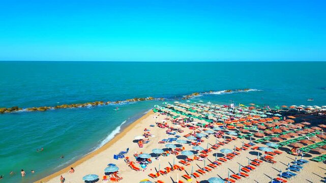 PORTO RECANATI, ITALY - JULY 30, 2025: Aerial rising push-in shot of the beach. Plenty of colored parasols and loungers lie on the beach of Porto Recanati, flooded with bathers in and out of the sea