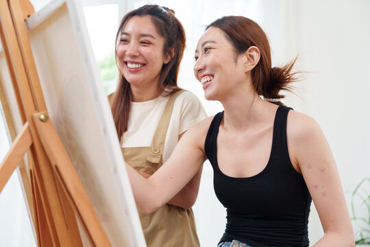 Two young beautiful, happy, cheerful Asian women smiling, laughing, painting watercolor together on canvas on wooden easel in cozy art studio at home with joy and fun