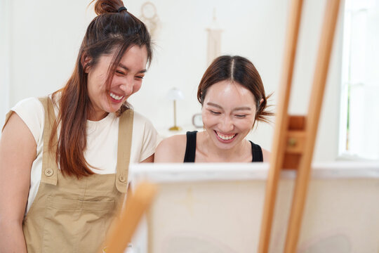 Two young beautiful, happy, cheerful Asian women smiling, laughing, painting watercolor together on canvas on wooden easel in cozy art studio at home with joy and fun. Friendship, mental wellbeing, di