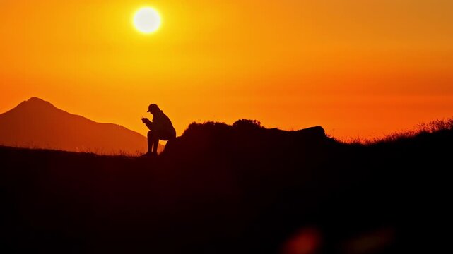 Solitary man sitting on a mountain summit using a smartphone against a beautiful orange sky in Col d'Aubisque in the French Pyrenees