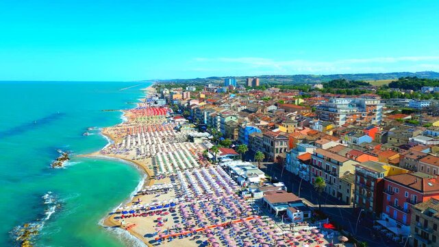 Revealing aerial pedestal up pullback shot from Porto Recanati, encompassing the Adriatic Sea waters splashing on the rocks and sand with white foam, the tourist-ready beach with equipment, buildings