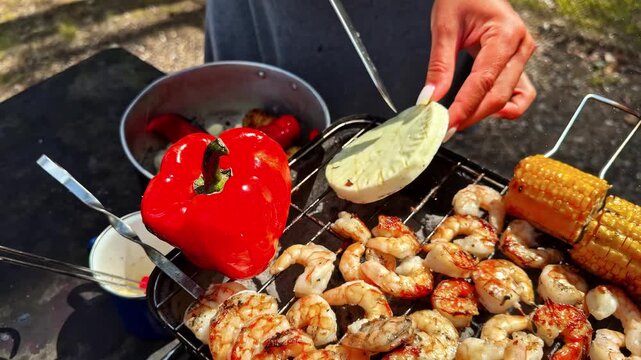 Hand places round piece of cheese onto grill beside grilled shrimp, corn on cob and red pepper, close-up of grilled lobster claw and pot of roasted vegetables