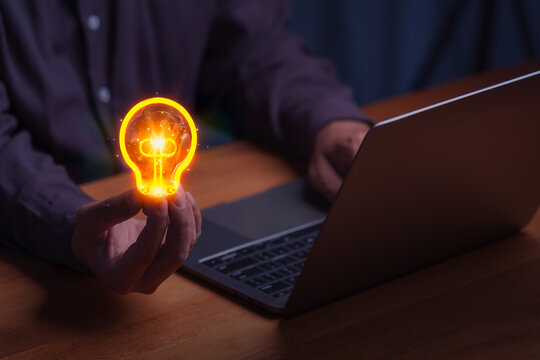 Adult man holds a glowing lightbulb over a laptop. This creative business concept represents digital technology innovation, offering a smart solution and inspiration for a new idea.