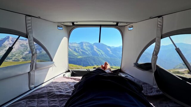 Person relaxes inside rooftop camping tent with panoramic view of green mountain range through window on sunny morning