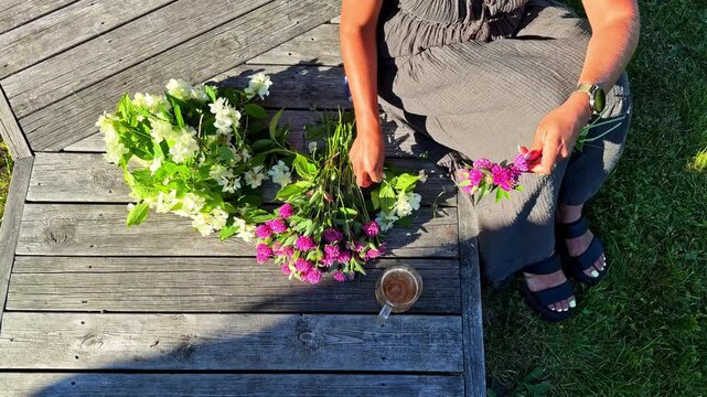 Woman weaving a flower crown with fresh jasmine and gomphrena on a wooden deck for Summer solstice in Latvia