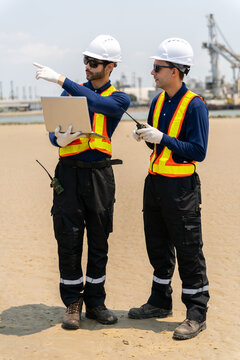 Two male engineers in safety vests and helmets discussing project details at shipyard port for logistics and industrial operations.