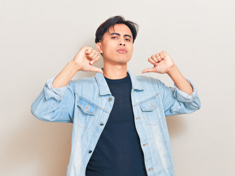 Young Asian man giving a double thumbs down gesture with an arrogant facial expression against a studio background while wearing a denim jacket.