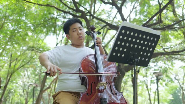Young musician practicing classical music outdoor. A teenage boy focusing on his cello performance with a music sheet stand in a peaceful garden concept for hobby, education, talent.