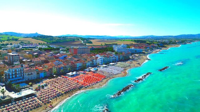 Sunny aerial push-in shot from Porto Recanati, going from above the Adriatic Sea with breakwaters to the sandy beach with parasols and resorts, lined with buildings, surrounded by hills, perfect sky