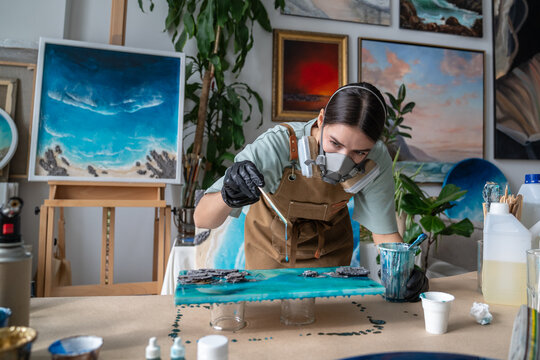 Concentrated woman handcraft master creating fluid art painting on canvas at worktable in studio, focused female artisan at workshop in respirator dripping liquid epoxy resin from stick over surface.