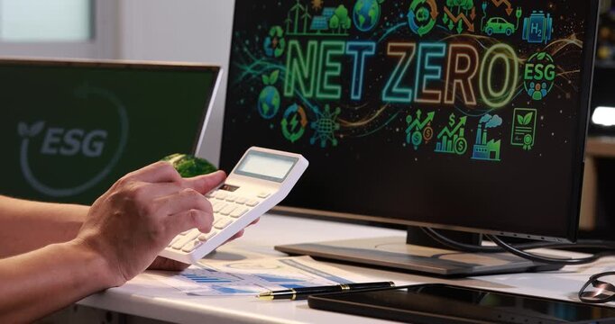 Hands using a calculator at an office desk with papers while a monitor shows net zero and esg graphics.