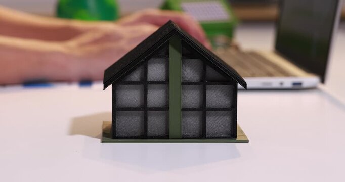 Small house model with dark front panels on a desk, with a blurred person and laptop in the background, suggesting home energy, building design, and sustainability.