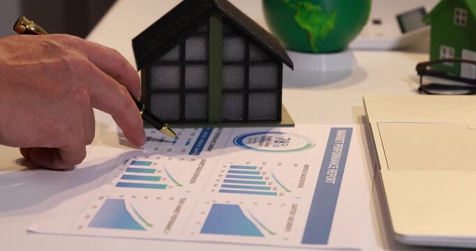 Hand pointing at a house model on a report with charts, energy efficiency, home planning, and green building ideas.