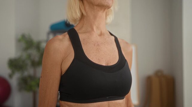 Woman in black bra tilts head back showing neck and chest in a yoga studio during gentle training with mat and exercise ball; calm strength.