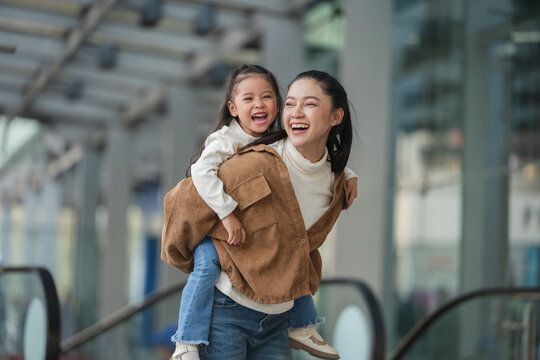 happy mother giving daughter piggyback ride near escalator in shopping mall or department store