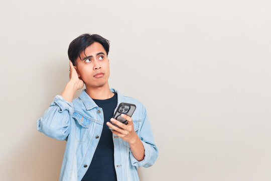 A confused young Asian man scratching his head and looking away against a neutral studio background while wearing a denim jacket