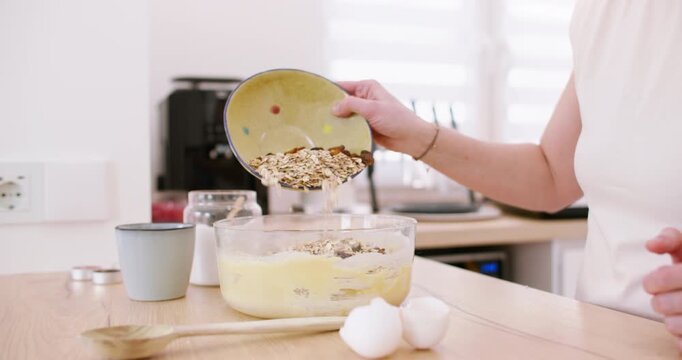 Homemade breakfast process. Healthy recipe. Female chef hand pouring oat flakes raisins in bowl with dough on modern kitchen counter.