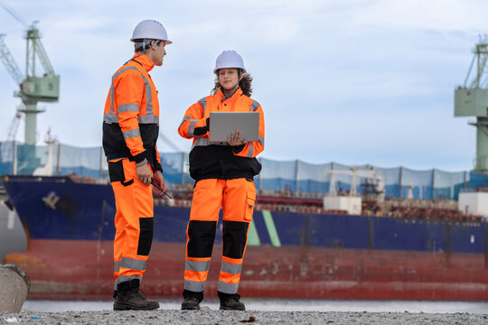 Logistic workers with laptop and radio standing at port shipping yard, Engineers in safety gear discussing cargo management by ship dock