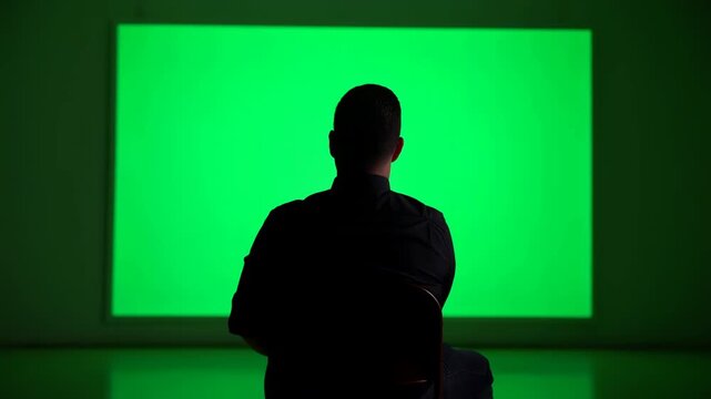 Rear silhouette of seated adult facing bright green panel with slight posture shift and arm movement inside dark studio setting, with silhouette, man, screen, green detail