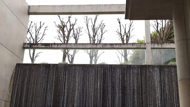 Wide view of contemporary architectural fountain with flowing water, raw concrete beams and leafless trees behind glass panels. Abstract urban background for design and travel themes. Kyoto, Japan.