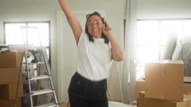 Woman raises arms beside cardboard boxes and a ladder inside a residential building during a move; moving day joy.